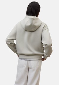 Persona con cabello oscuro, vistiendo una sudadera holgada de color gris claro y pantalones blancos, de pie con las manos en las caderas, de espaldas a la cámara.