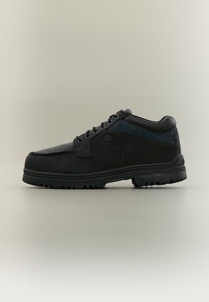 Chaussure noire basse à lacets avec semelle robuste et col rembourré, logo visible sur le côté, fonds neutre.