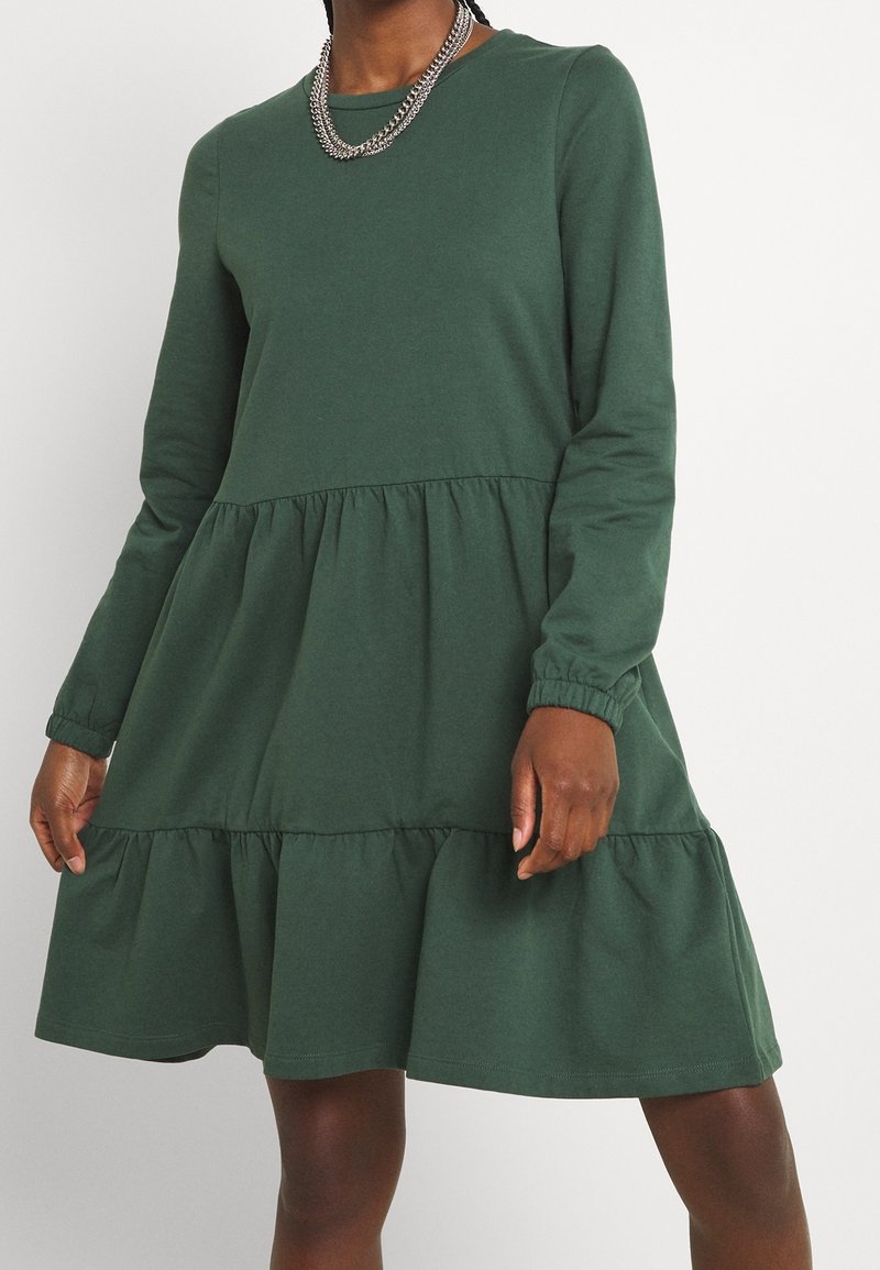 Robe en coton vert avec des manches longues, un col rond et un ourlet à volants en couches. Comprend des poignets élastiques et une texture douce et lisse.