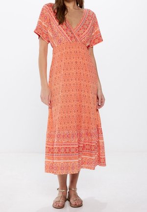Femme portant une robe midi orange à motifs avec un décolleté en V, manches courtes, et des sandales plates beige à brides, sur un fond blanc.