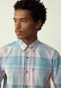 Camicia azzurro chiaro a quadri con strisce rosa pastello e avorio, dotata di colletto appuntito e bottoni frontali. Include un piccolo taschino.