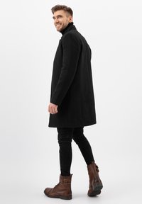 Cappotto in lana nera con colletto alto, design elegante, tasche laterali, abbinato a stivali in pelle marrone con dettagli in fibbia e suole testurizzate.