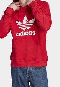 Osoba upravující kapuci červené mikiny Adidas s bílým logem, oblečená v modrých džínách, na jednobarevném pozadí.