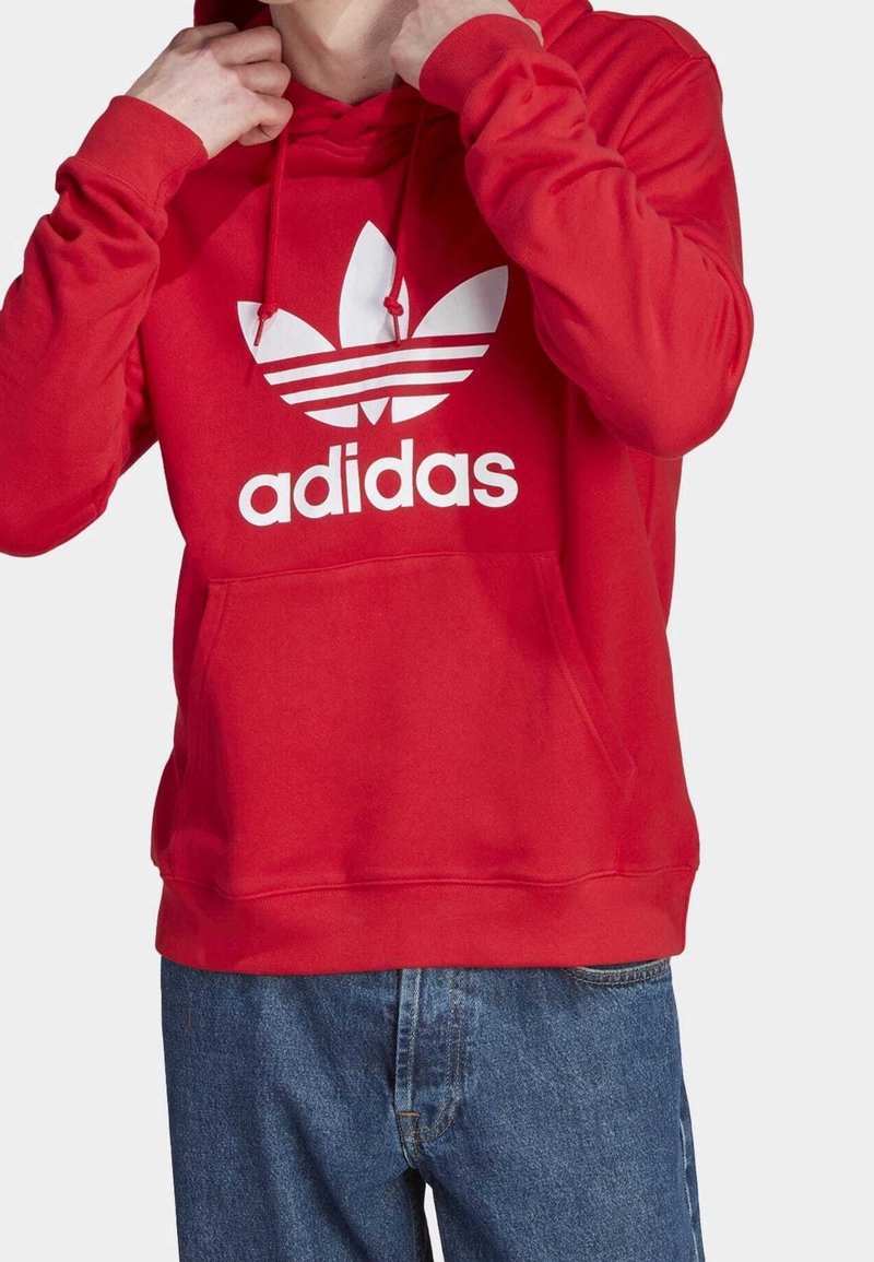 Osoba upravující kapuci červené mikiny Adidas s bílým logem, oblečená v modrých džínách, na jednobarevném pozadí.