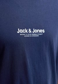 Tissu bleu marine avec texte blanc sur le devant indiquant "Jack & Jones, Brought to you by Originals Studio, Designed in Copenhagen".