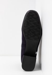 Semelle d'une chaussure noire à bout carré avec un talon bloc, présentée debout contre un mur et un sol blancs.