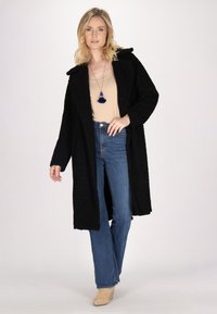 Schwarzer, strukturierter Überwurf mit Kragen, getragen über einem beigen Oberteil und blauen Jeans. Das Modell ist mit einer blauen Quasten-Halskette accessoriert.