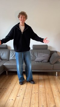 Giacca nera in pile con zip, camicia bianca sotto, jeans larghi blu e scarpe nere, in piedi su pavimento in legno con divano grigio alle spalle.