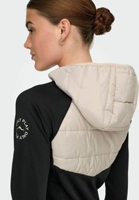 Črna in svetlo rjava jakna z napihnjenim kapuco, s kontrastnim blokiranim dizajnom in vezenim napisom na rokavu.