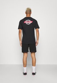Camiseta negra de manga corta con un logo "Dickies" en rojo y blanco en la parte de atrás, combinada con pantalones cortos negros y zapatillas con un diseño.