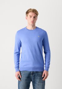Helles blaues Strickpullover mit Rundhalsausschnitt und langen Ärmeln, mit einem kleinen gelben Logo auf der Brust. Getragen mit blauen Jeans.