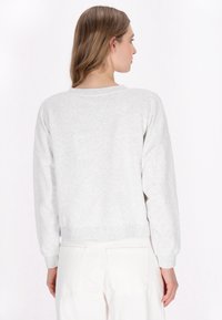 Sweatshirt gris clair avec une coupe décontractée, des bords côtelés aux poignets et à l'ourlet ; tissu doux et texturé ; couture visible ; porté avec un jean blanc.