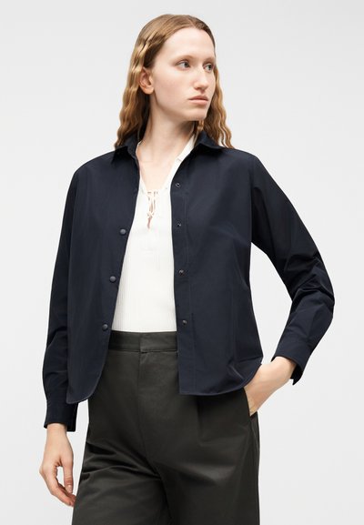 Femme aux longs cheveux ondulés portant une chemise bleu marine boutonnée sur un haut blanc et un pantalon noir, debout, une main dans la poche, regardant vers la droite.