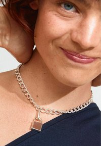 Choker en chaîne argentée présentant un pendentif en forme de cadenas, avec des maillons lisses et une finition polie ; porté avec un haut bleu.