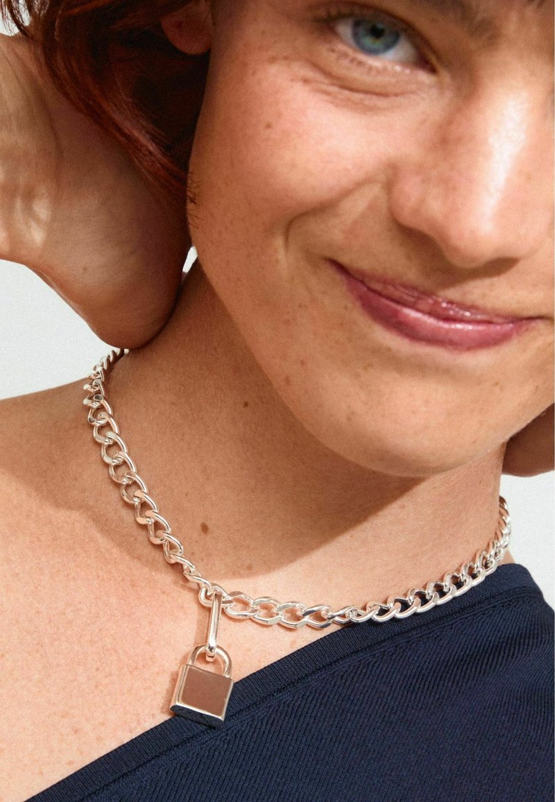 Srebrna choker ogrlica sa privjeskom u obliku brava, s glatkim karikama i poliranim završetkom; nosi se uz plavu majicu.