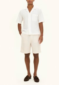 White short-sleeve shirt with a pocket, paired with light beige shorts. The outfit features a relaxed design and soft texture. Brown loafers complete the look.