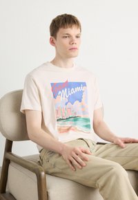 Helebeige puuvillane T-särk, millel on värvikas Miami graafika, lühikesed varrukad ja ümar kaelus. Mudel istub puidust toolil.
