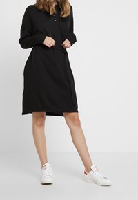 Robe noire à manches longues avec un col et des boutons sur le devant, dotée d'une taille ceinturée et poches latérales. Portée avec des baskets blanches.