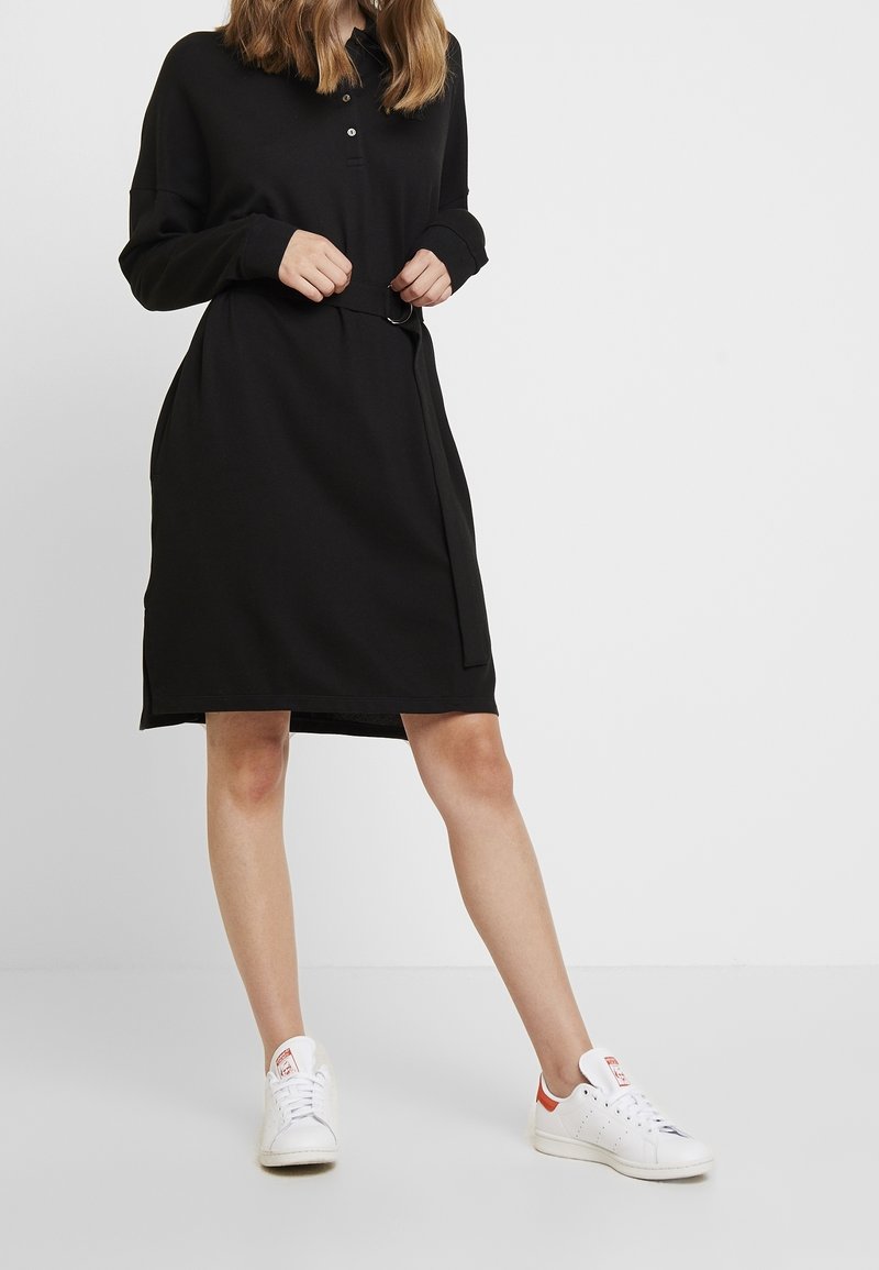 Robe noire à manches longues avec un col et des boutons sur le devant, dotée d'une taille ceinturée et poches latérales. Portée avec des baskets blanches.