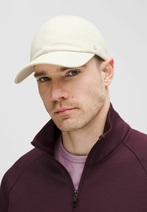 Gorra blanca con visera curva, hecha de tela suave. Llevada con una chaqueta de cremallera morada oscura y una camisa morada clara debajo.