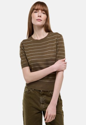 Femme aux cheveux roux lisses portant un pull rayé à manches courtes vert olive et un pantalon assorti vert olive, sur fond blanc.