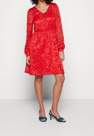 Kvinde iført en rød, knælængde kjole med lange ærmer i blonder og V-hals samt turkise højhælede sandaler med åben tå, stående foran en ensfarvet baggrund.