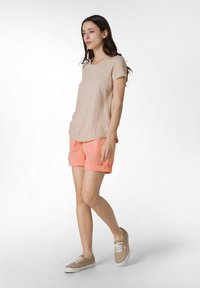 Beige kortärmad topp med en mjuk textur, kombinerad med ljus korallfärgade shorts med uppvikta ärmar. Modellen bär beige snörskor.
