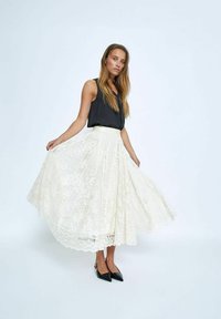 Weiße Spitzen-Midi-Rock mit filigranen Blumenmustern, kombiniert mit einem schwarzen ärmellosen Oberteil und spitzen schwarzen Ballerinas, vor einem hellen Hintergrund präsentiert.