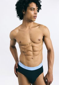 Fekete pamut alsónadrág világoskék derékszíjjal, amelyen a "Calvin Klein" logó látható. A modell egy sima fehér háttér előtt van megformázva.