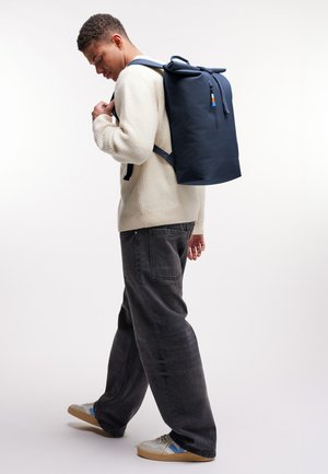 Young man adjusting straps of large navy backpack, wearing white sweater, black wide-leg pants, and white sneakers with blue accents.