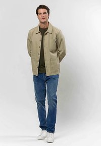 Homme debout, les mains dans le dos, portant une veste beige boutonnée, une chemise vert foncé, un jean bleu et des baskets blanches sur un fond uni.
