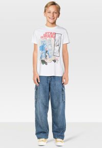 Camiseta blanca con un gráfico de "Star Wars", que presenta ilustraciones coloridas al estilo cómic. Pantalones de cargo de mezclilla azul sueltos con bolsillos laterales. Zapatillas amarillas.