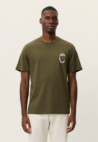 Βαμβακερό μπλουζάκι σε ελαιί πράσινο με στρογγυλή λαιμόκοψη και κοντά μανίκια. Διαθέτει διακοσμητικό τσιμπιδάκι στο αριστερό στήθος με γραφικό σχέδιο.