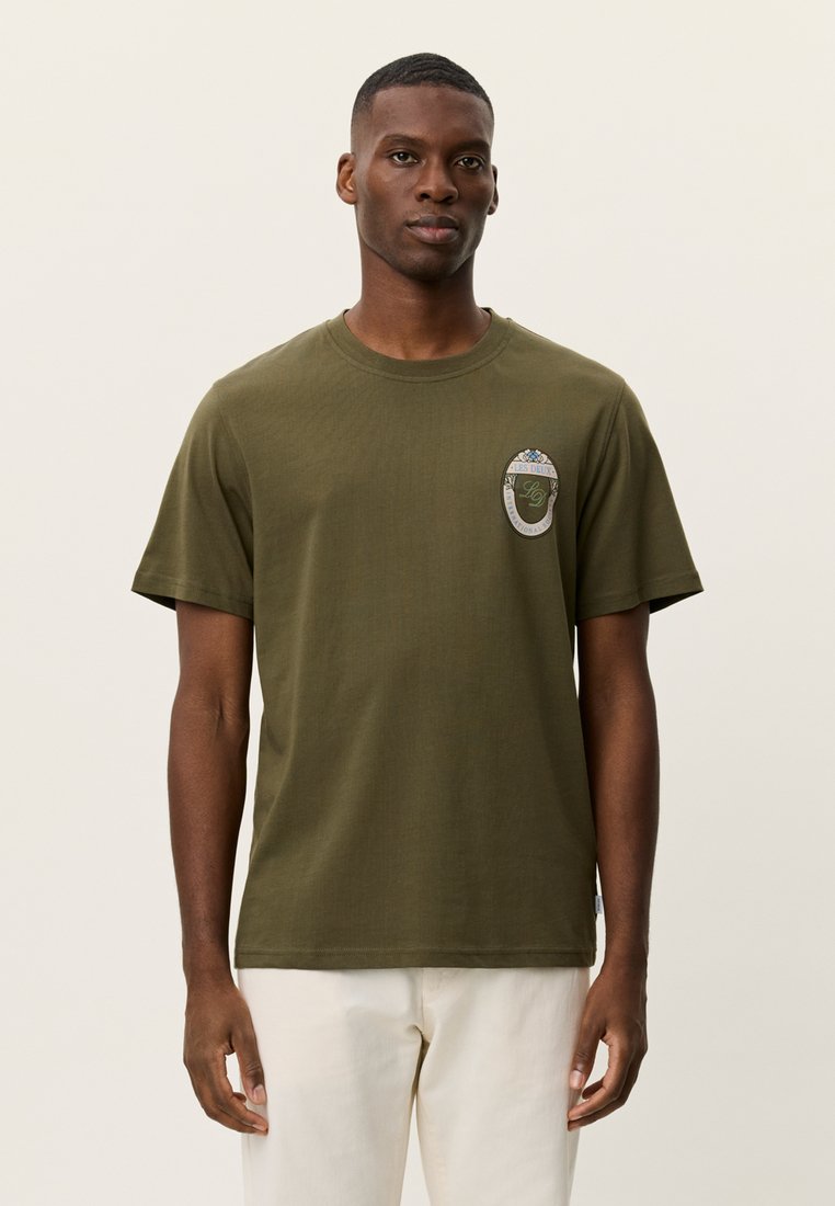 Βαμβακερό μπλουζάκι σε ελαιί πράσινο με στρογγυλή λαιμόκοψη και κοντά μανίκια. Διαθέτει διακοσμητικό τσιμπιδάκι στο αριστερό στήθος με γραφικό σχέδιο.