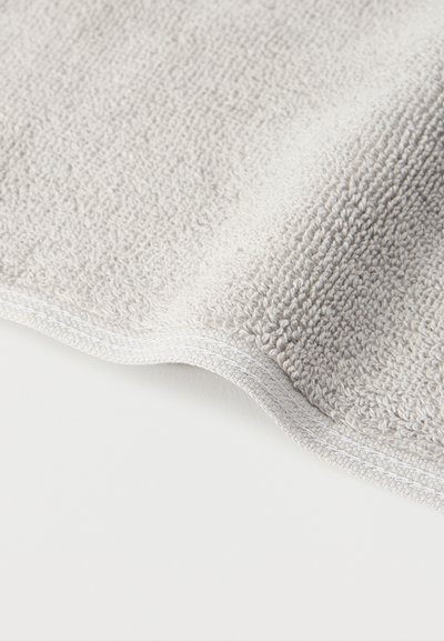 Υφασμα πετσέτας από ελαφρύ γκρι ύφασμα με ορατές θηλιές και περιμετρικό ραφή σε λευκό φόντο.