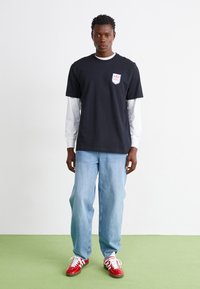 Black cotton t-shirt layered over a white long-sleeve shirt, paired with light blue denim jeans and red sneakers, with a logo patch on the chest.