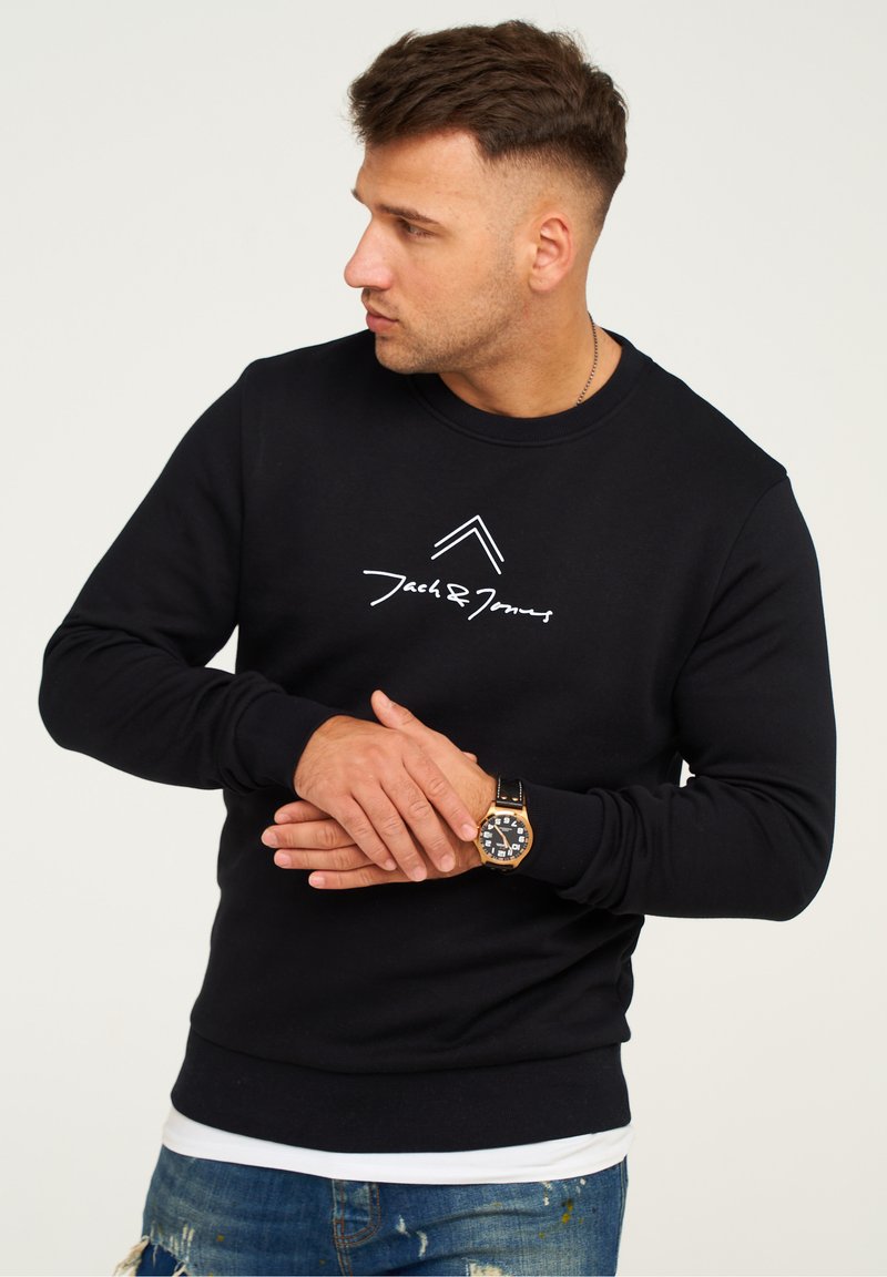 Black sweatshirt with crew neckline, featuring white logo print. The model wears distressed blue jeans and a black watch.