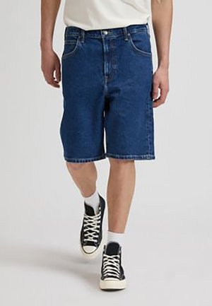 Hombre que lleva pantalones cortos de mezclilla azul oscuro, calcetines blancos, zapatillas negras altas y una camisa blanca caminando hacia adelante sobre un fondo liso.