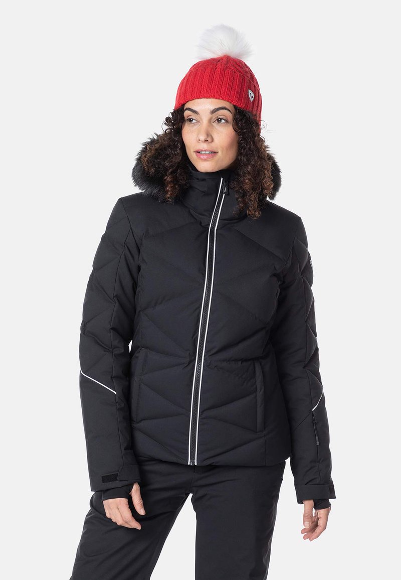 Svart puffjacka med quiltat mönster, dragkedja fram och vita accentlinjer; har en foder med päls på huvan. Modellen bär en röd stickad mössa.