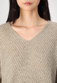 Femme portant un pull en maille beige texturé avec un col en V, visible du cou au milieu du torse sur un fond uni.