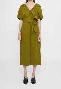 Olivově zelené šaty s výstřihem do V, nafouknutými rukávy, uvázaným pasem a širokými zkrácenými nohama. Nosené s černými botami na minimalistickém pozadí.