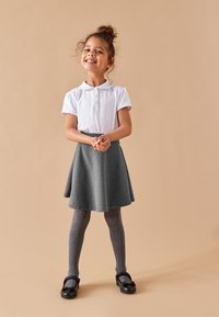 Valkoinen lyhythihainen paita, jossa on kaulus, harmaa levenevä hame, harmaat sukkahousut ja mustat kengät, joissa on rusetti. Yksinkertainen tausta korostaa asun yksityiskohtia.