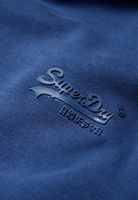 Modrá tkanina s vyraženým textem "SuperDry" a stylizovaným podtržením, s hladkou texturou a čistým designem.