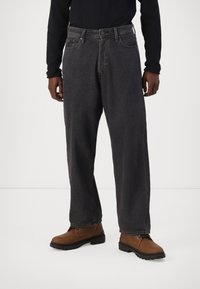 Jeans negri din denim cu o croială relaxată, având un design clasic cu cinci buzunare și cusături contrastante, asortați cu cizme de muncă maro.