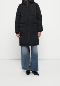 Sort polstret parka med hætte, to forlommer, lynlås og ribstrikkede manchetter. Kombineret med vidbenede, falmede blå jeans og sorte hæle.