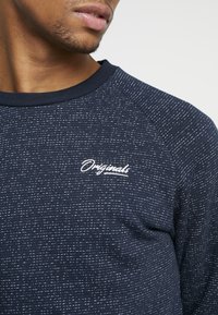 Man som bär en marinblå, prickentröja med vit "Originals" broderad på bröstet, visar nedre delen av ansiktet och nacken.