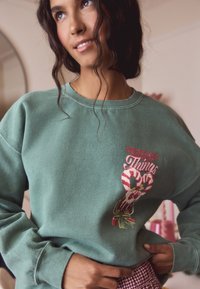 Žalias džemperis su siuvinėta saldainių lazdelės schema ir užrašu "SEASONS GREETINGS." Minkšta tekstūra ir laisvas kirpimas su riešinėmis.