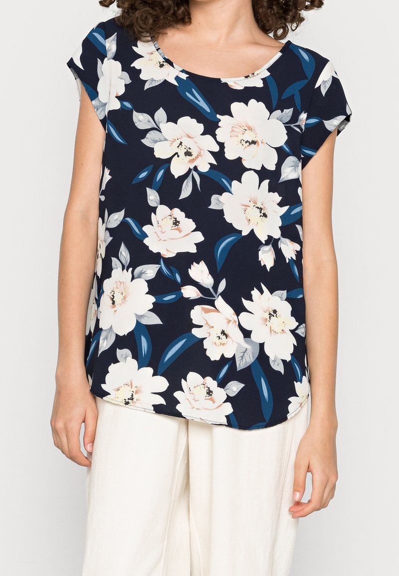 Haut floral en tissu bleu marine avec des motifs de fleurs blanches, roses et vertes. Manches courtes et encolure ronde. Conception ample.