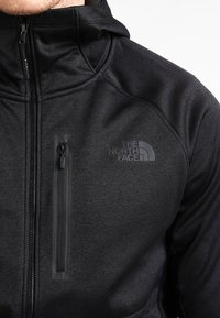 Zwarte zip-up jas met een textuurstof, voorzien van een met rits sluitend borstzakje en een logo aan de linkerkant. Met capuchon.