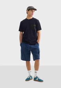 Černé bavlněné tričko s kapsou, modré cargo šortky s bočními kapsami, šedá kšiltovka, bílé ponožky, modré tenisky s bílými akcenty.